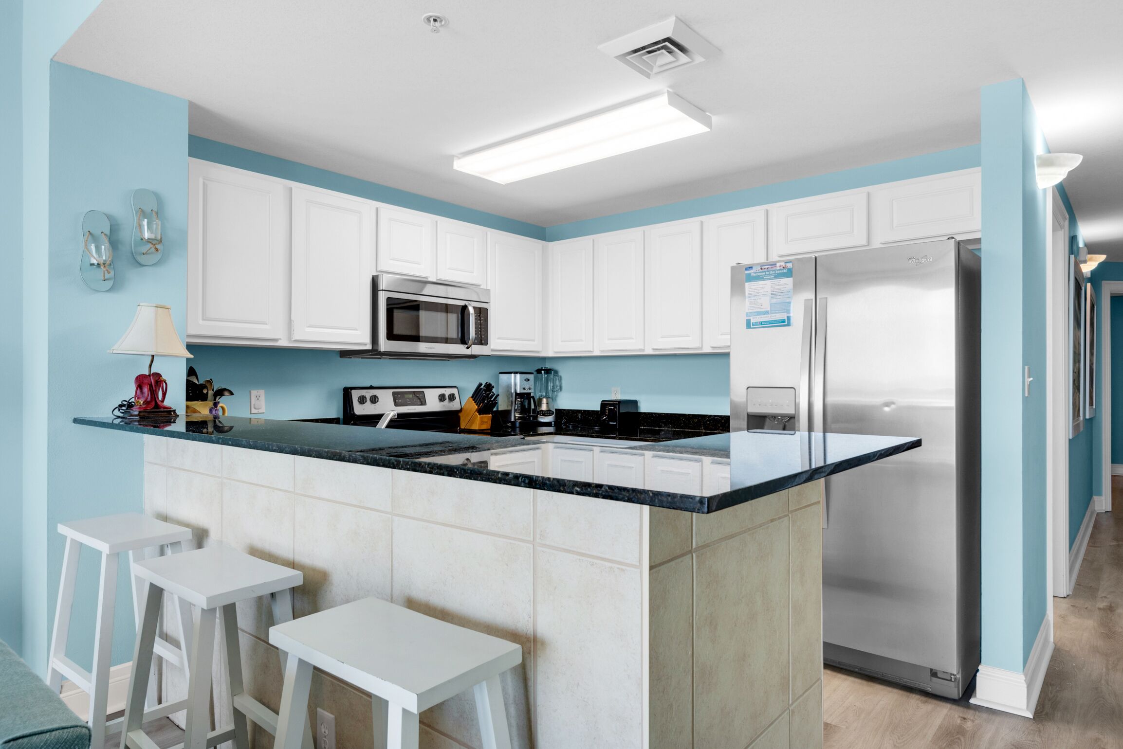 A modern kitchen with white cabinets, a stainless steel refrigerator, a microwave, and a black countertop. There are two white bar stools and a small table.