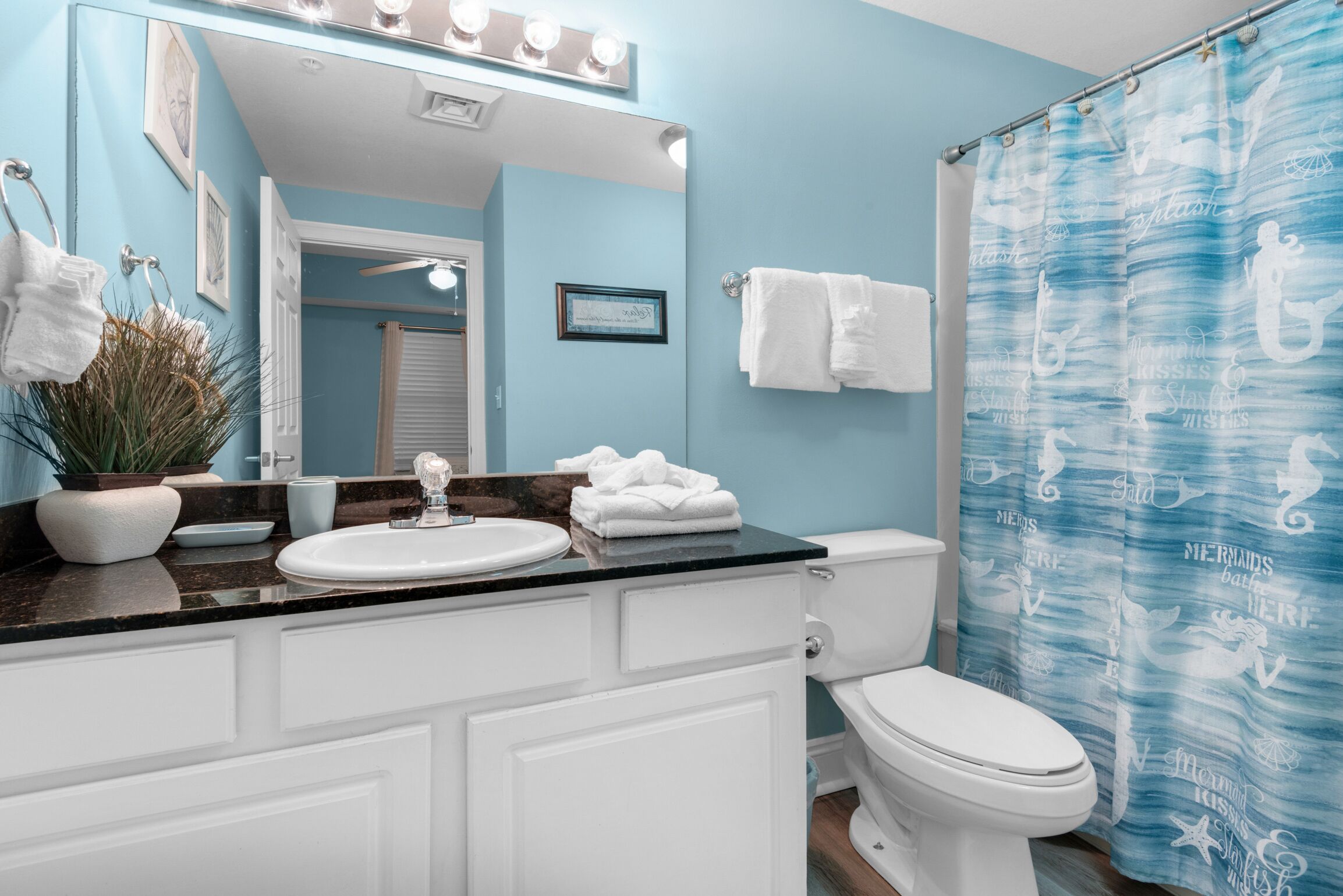 A bathroom with a white sink, a toilet, and a shower curtain with a mermaid theme. There are towels hanging on the wall and a potted plant on the counter.