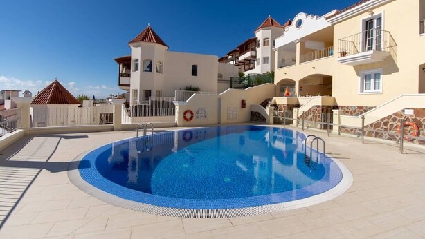 A swimming pool surrounded by buildings with balconies, a lifebuoy, and a clear sky. - Bahia la Caleta 1 - Two Bed (La Caleta)
