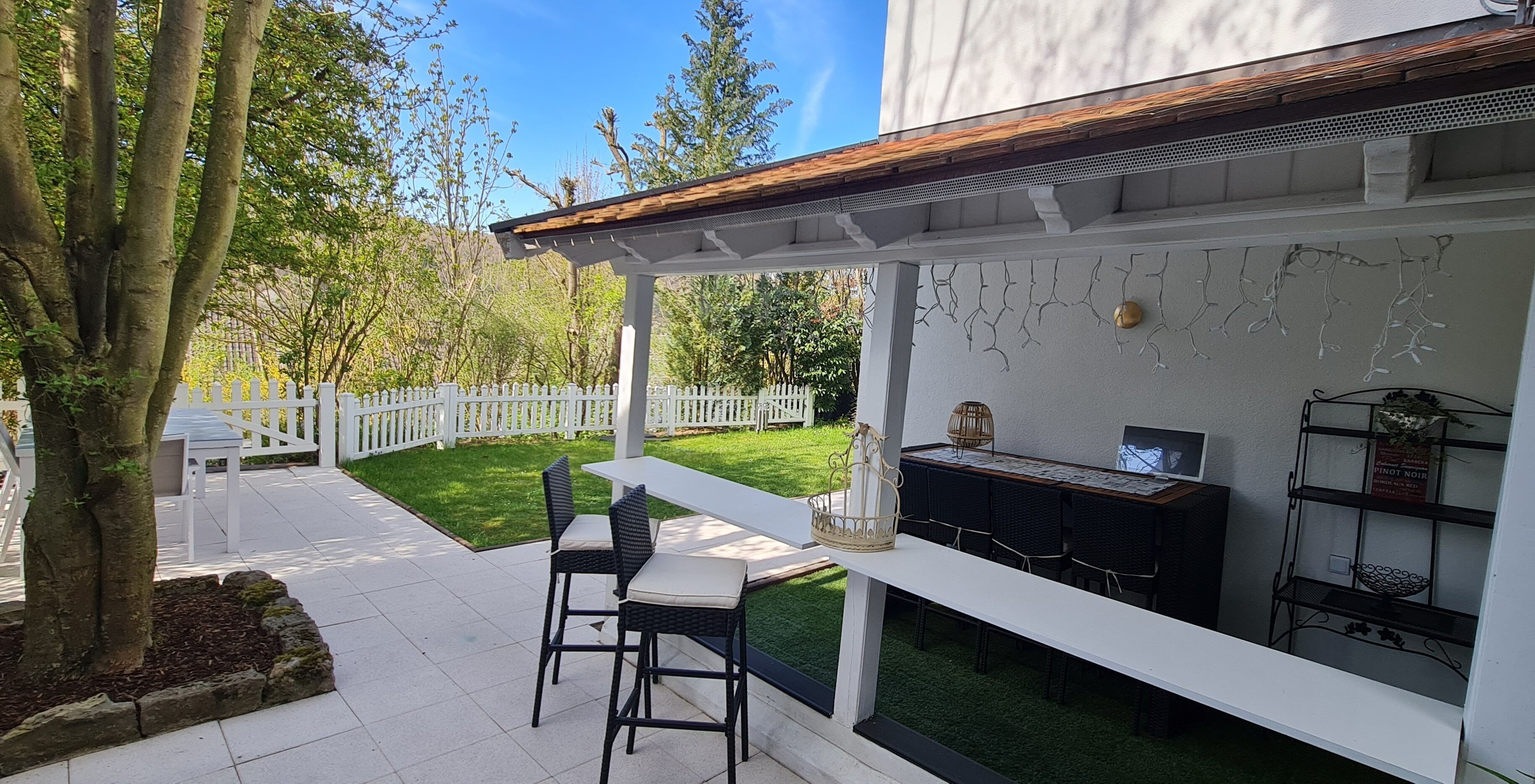 An outdoor patio with a white pergola, a dining table with chairs, a grill, and a white fence in the background.
