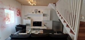 A modern living room with a flat-screen TV, black leather sofas, a glass coffee table, and a staircase with a white railing.