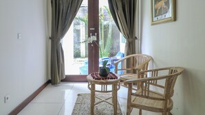 Una habitación con una silla de madera y una mesita con un florero y una planta. Se ve una puerta que da a un espacio exterior con una silla azul y una planta.