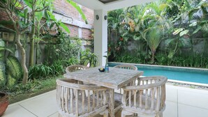 Un patio exterior con mesa y sillas de madera, rodeado de exuberante vegetación tropical y vista a una piscina.