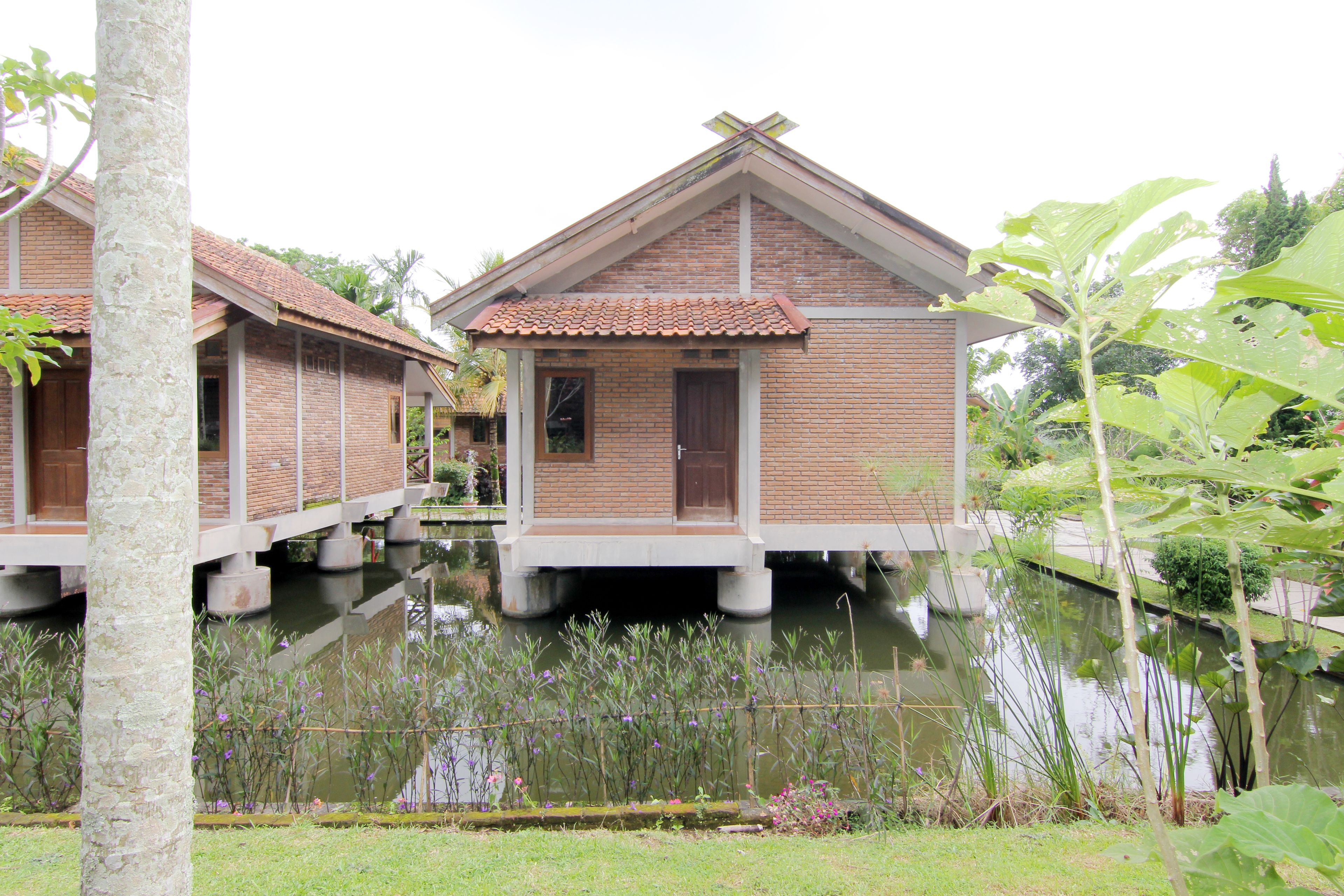 Two wooden overwater bungalows with brick walls and tiled roofs, floating on a calm water body, surrounded by lush greenery.