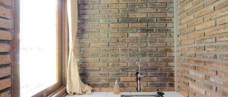 Una cocina con pared de ladrillo, una ventana con cortinas, un lavabo y un dispensador de jabón.