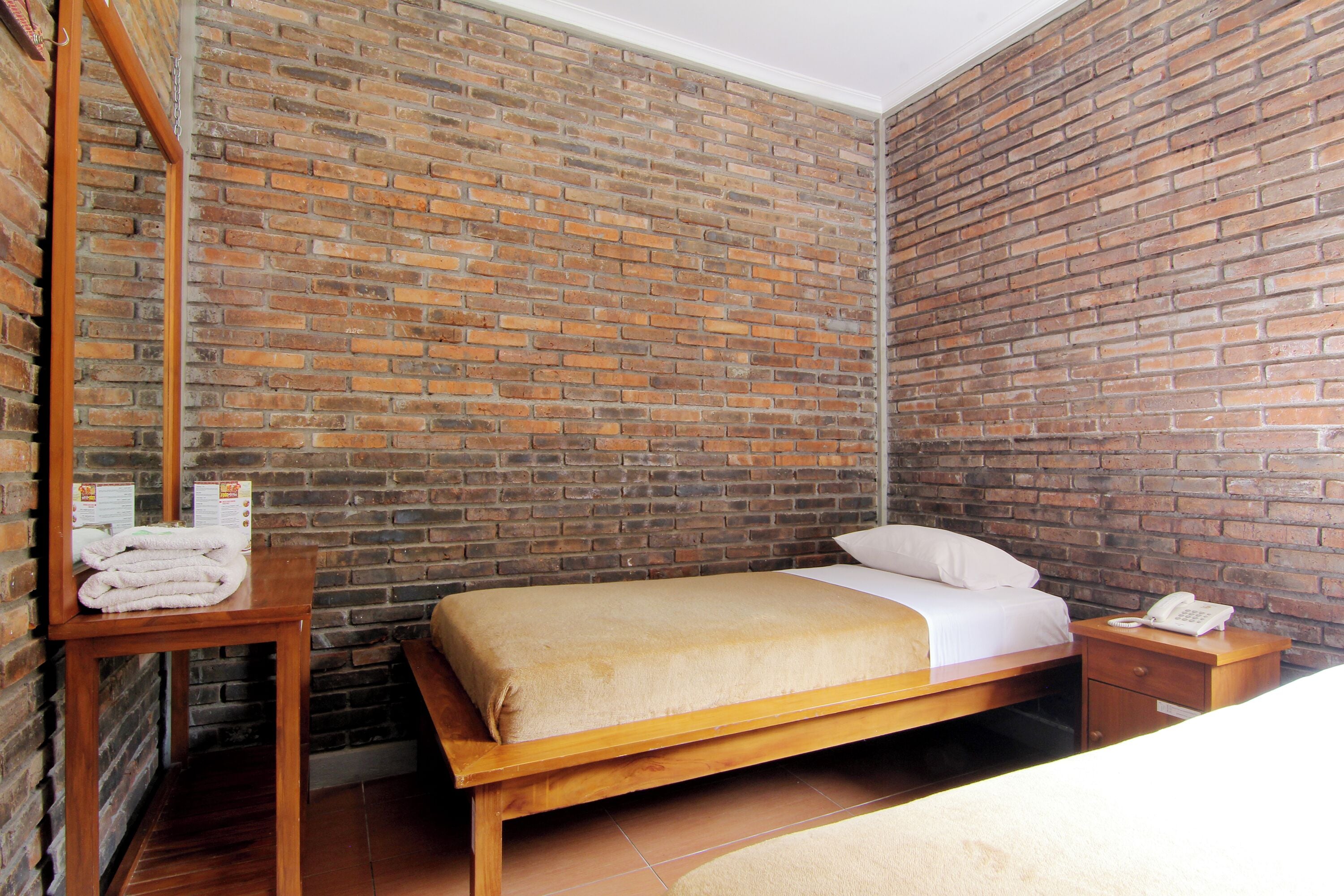A hotel room with a brick wall, a wooden bed, a bedside table with a telephone, and a small table with towels.