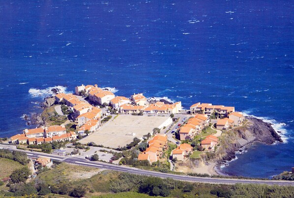 An aerial view of a coastal resort with multiple buildings, a central open area, and a road leading to the shore.