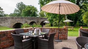 Un espace extérieur aménagé en terrasse, avec des meubles en osier et une table dressée pour les repas, le tout entouré de murs en briques et de verdure.