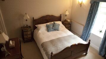 A neatly made bed with pillows, a bedside table with a lamp, a nightstand with a lamp, and a window with curtains.