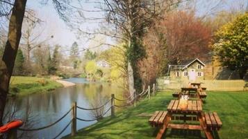 A lakeside picnic area with a wooden table and benches, surrounded by a fence and trees.