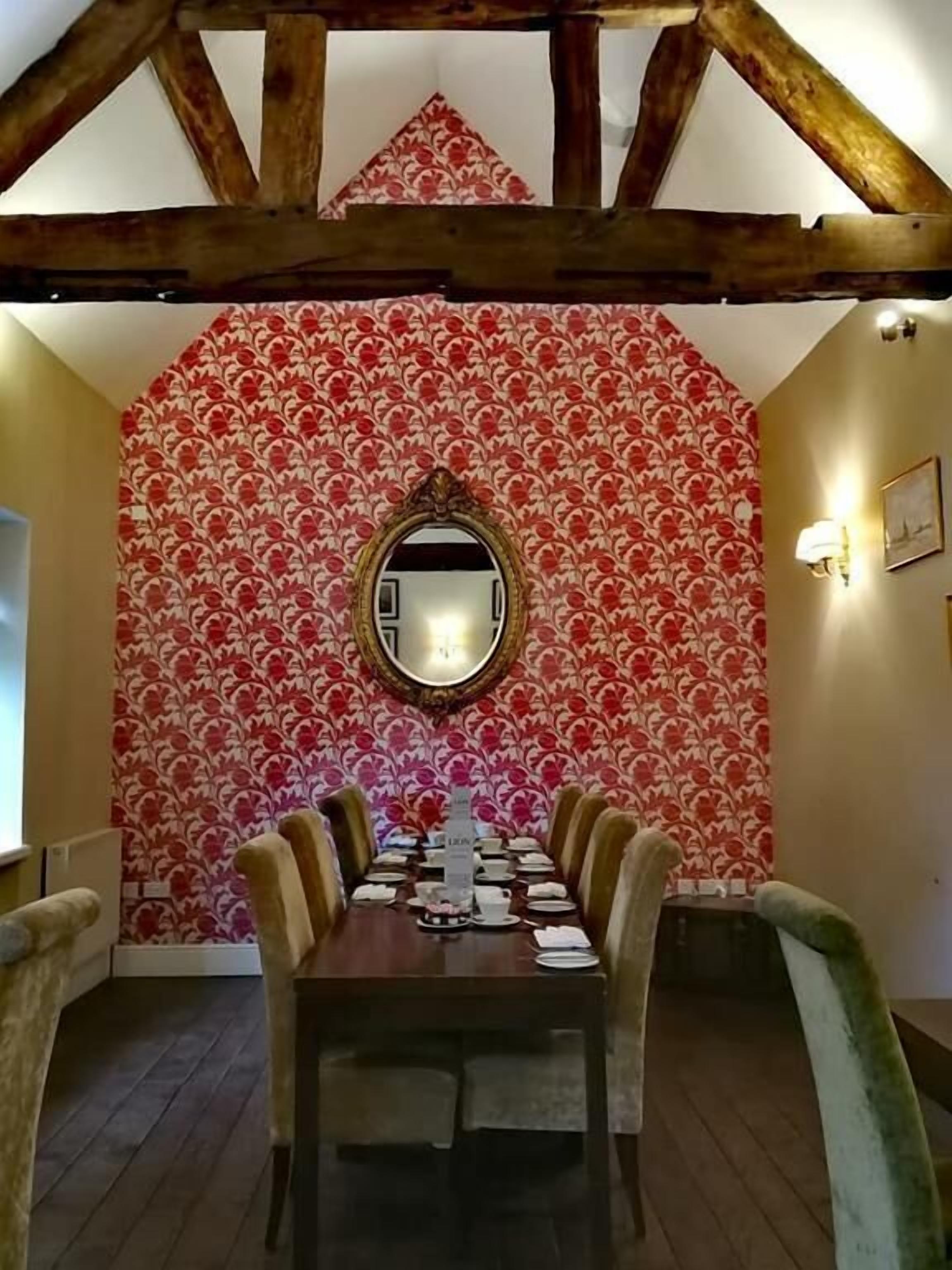 Comedor con mesa de madera preparada para seis comensales, papel tapiz estampado y un espejo sobre la mesa.