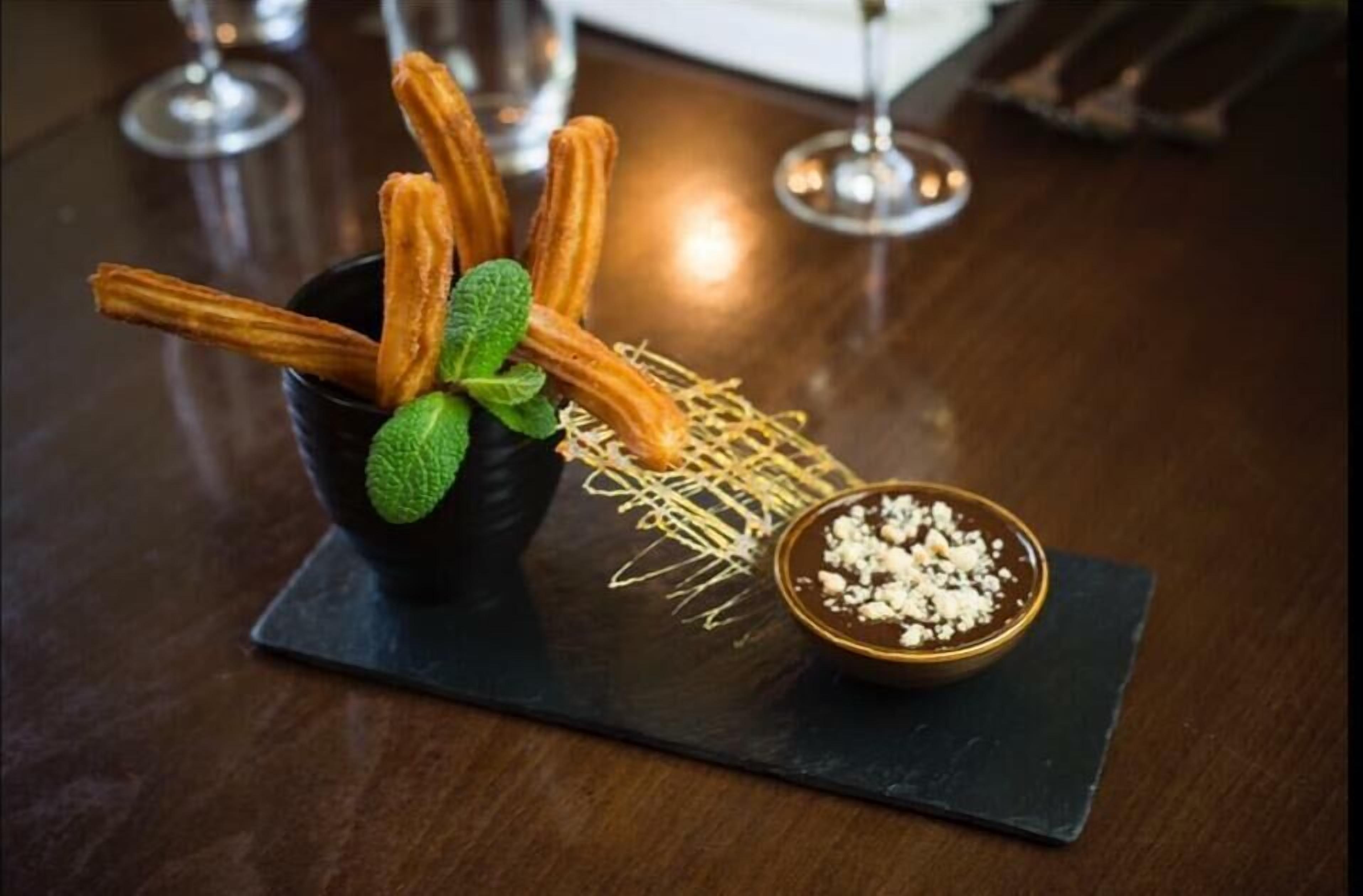 Un plat de postres amb xurros, una fulla de menta i un bol de salsa de xocolata sobre una placa de pedra.