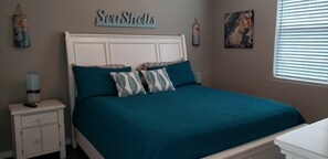 Quarto com colcha azul-turquesa, cabeceira branca e almofadas a combinar. Há uma mesa de cabeceira com abajur e uma gaveta pequena. Na parede, um letreiro com a inscrição 'SeaShells' e duas obras de arte emolduradas.