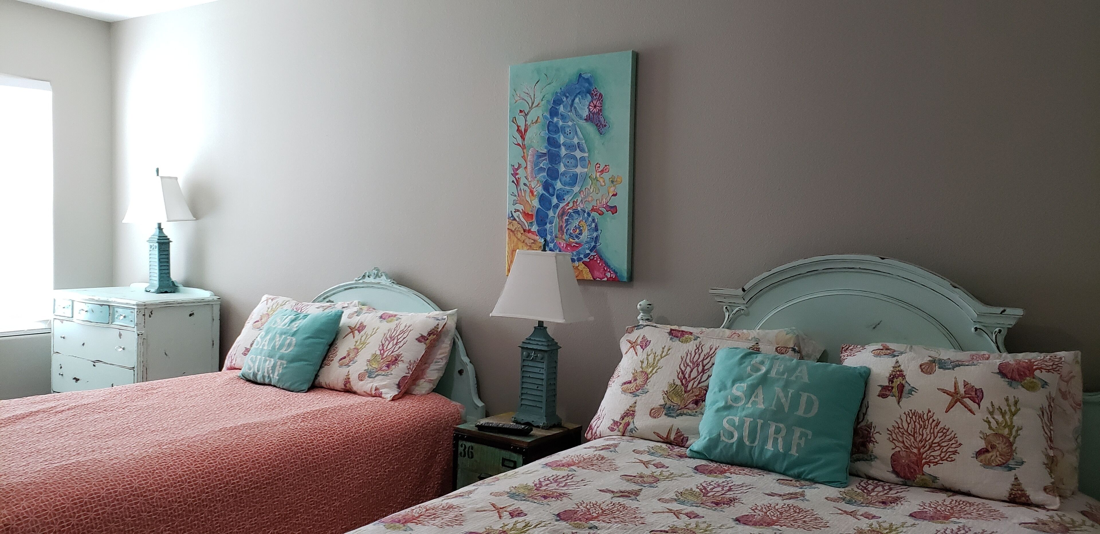 Une chambre avec deux lits, chacun agrémenté de draps et d’oreillers à motifs floraux assortis. Un tableau coloré représentant un hippocampe est accroché au mur, au-dessus des lits.