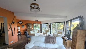 A spacious living room with a white sofa, two armchairs, a wooden cabinet, and a large window offering a view of trees.