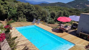 A rectangular swimming pool with a wooden deck, surrounded by lush greenery and hills in the background.