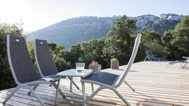 An outdoor deck with two lounge chairs, a small table, and a glass of water. The deck overlooks a hillside with buildings and greenery.