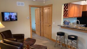 A living room with a TV, leather chairs, a coffee table, and a kitchen area with wooden cabinets and a countertop. - Park Right by Your Door -1 BR, 1 Bath, Full Kitchen, 1st Floor Laundry. (Lansing)