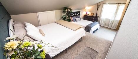 Un dormitorio con dos camas, una mesita de noche con lámpara, una planta en maceta y una ventana con cortinas.