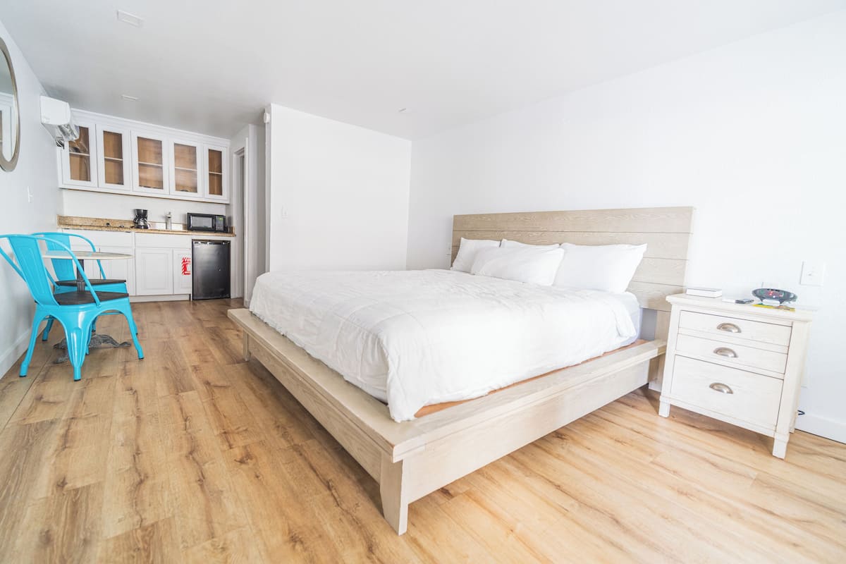 A bedroom with a wooden bed, a nightstand, a blue chair, a kitchen area with cabinets, and a microwave.