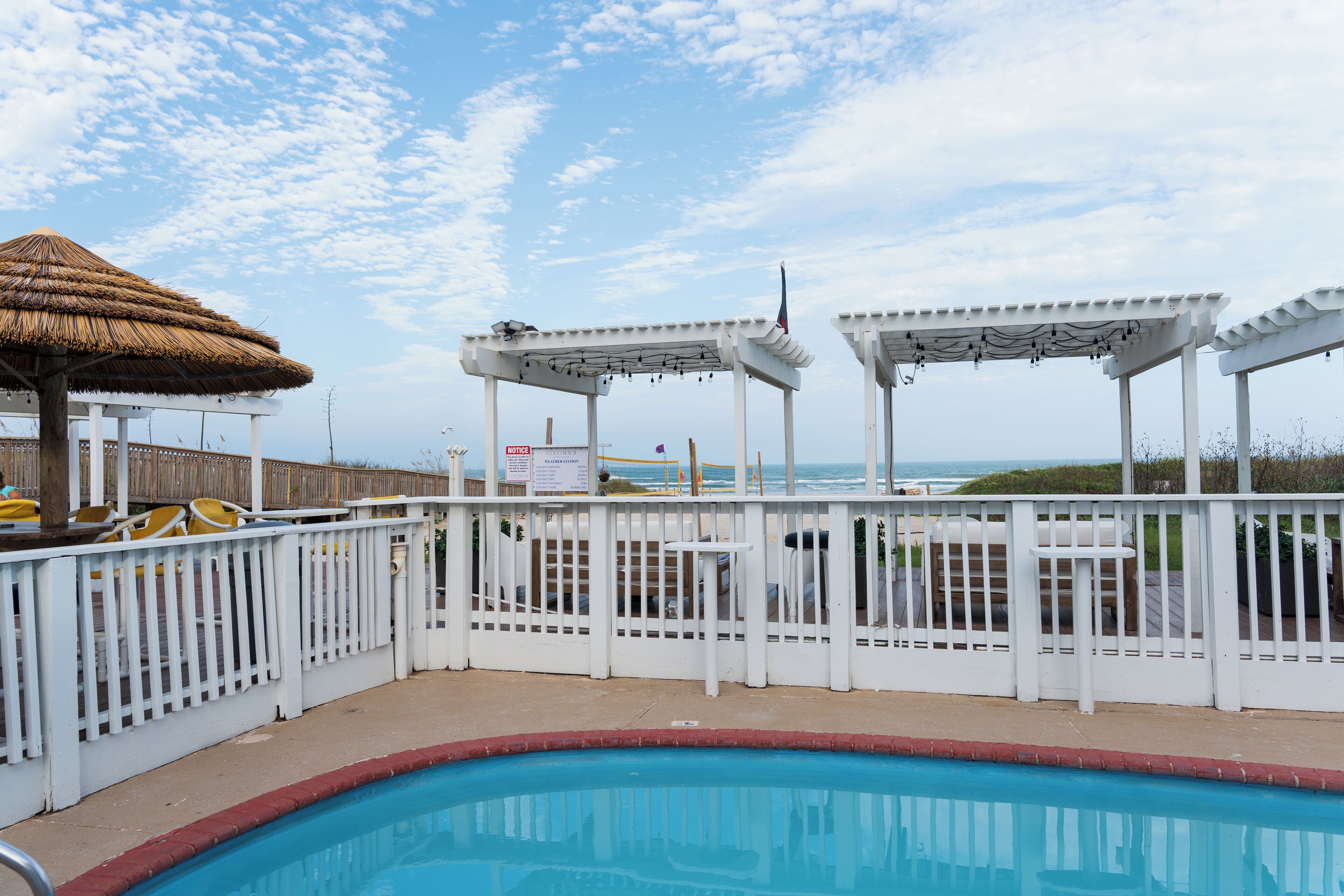A pool area with a thatched umbrella, white pergolas, and a view of the ocean.