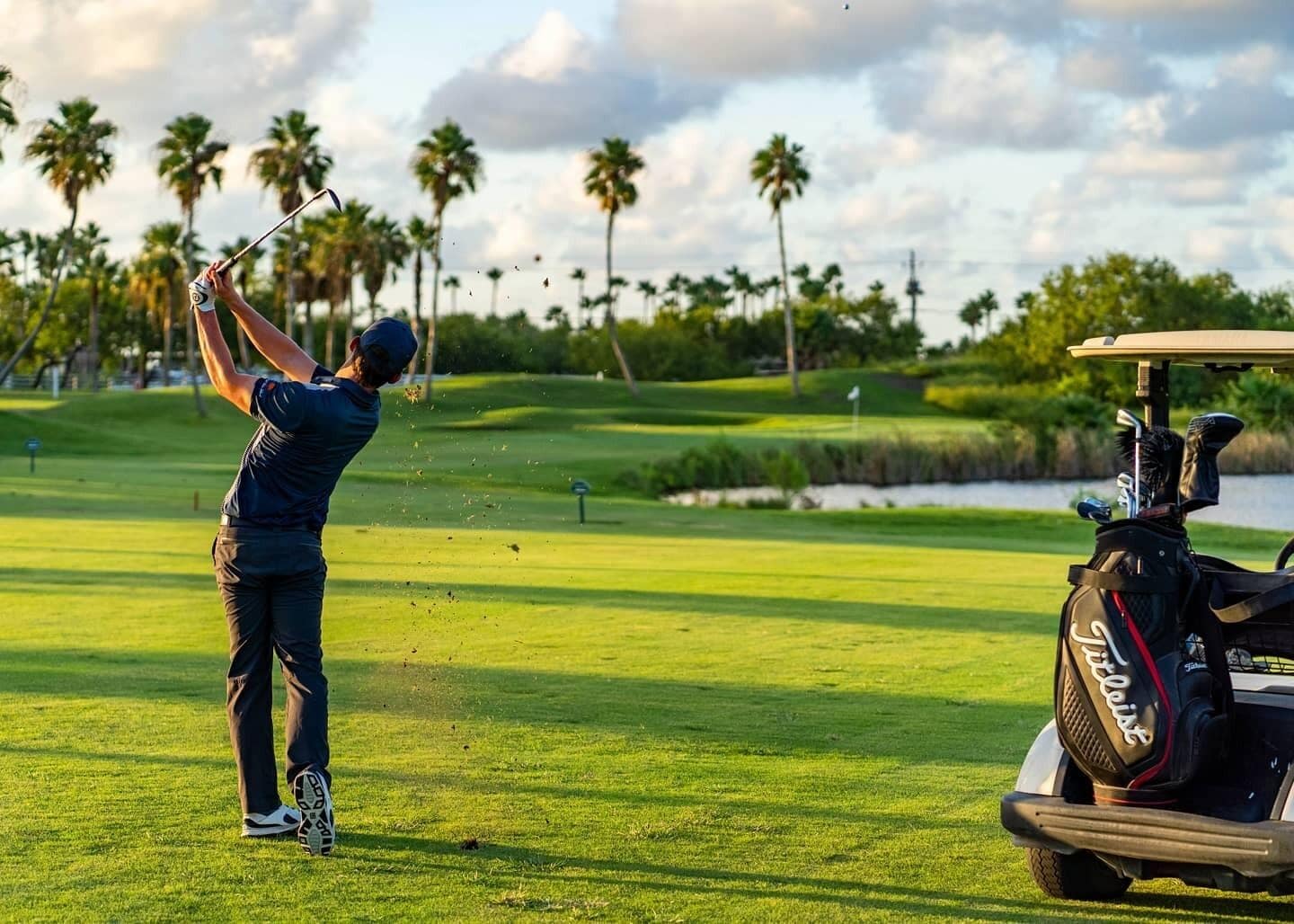 A golf course with a player in mid-swing, palm trees, and a golf cart in the background.