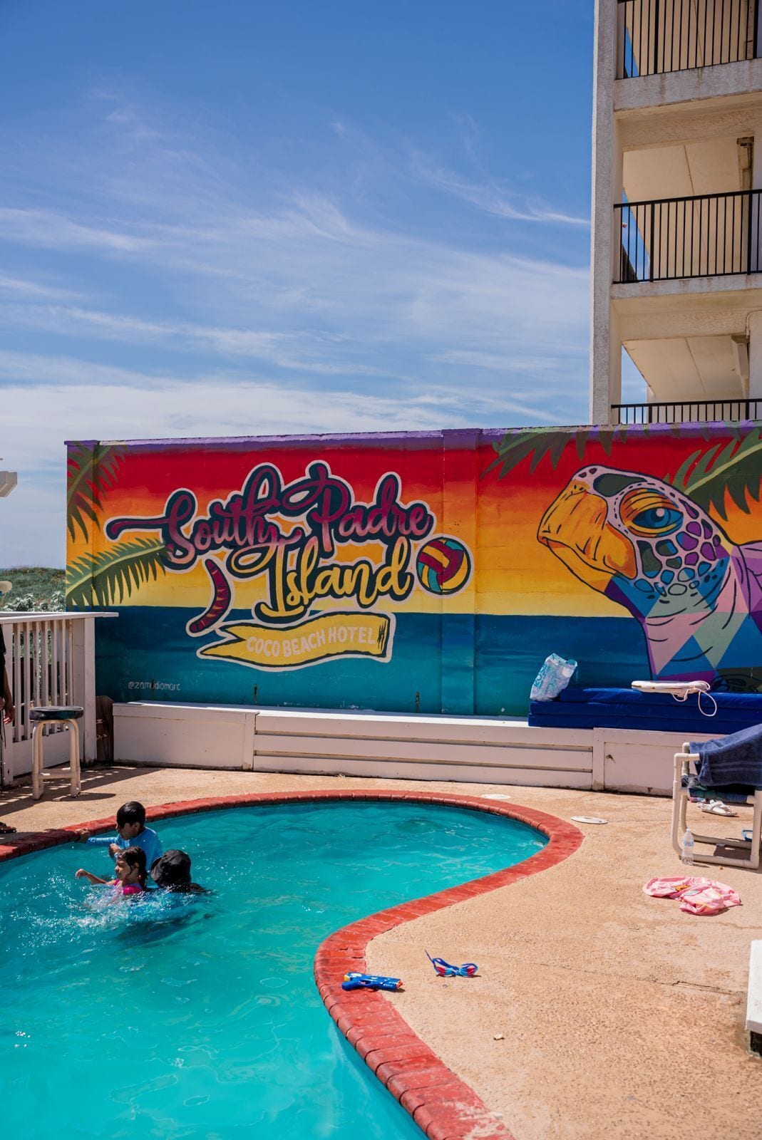 Un mural colorido con un tucán y palmeras en una pared de hotel, un área de piscina con borde rojo y una silla de salvavidas.
