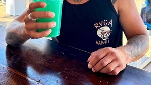 A person sitting at an outdoor table with a drink, wearing a black tank top with a logo, and surrounded by a sunny outdoor setting with umbrellas and chairs. - Coco Beach Hotel on South Padre Island (South Padre Island)