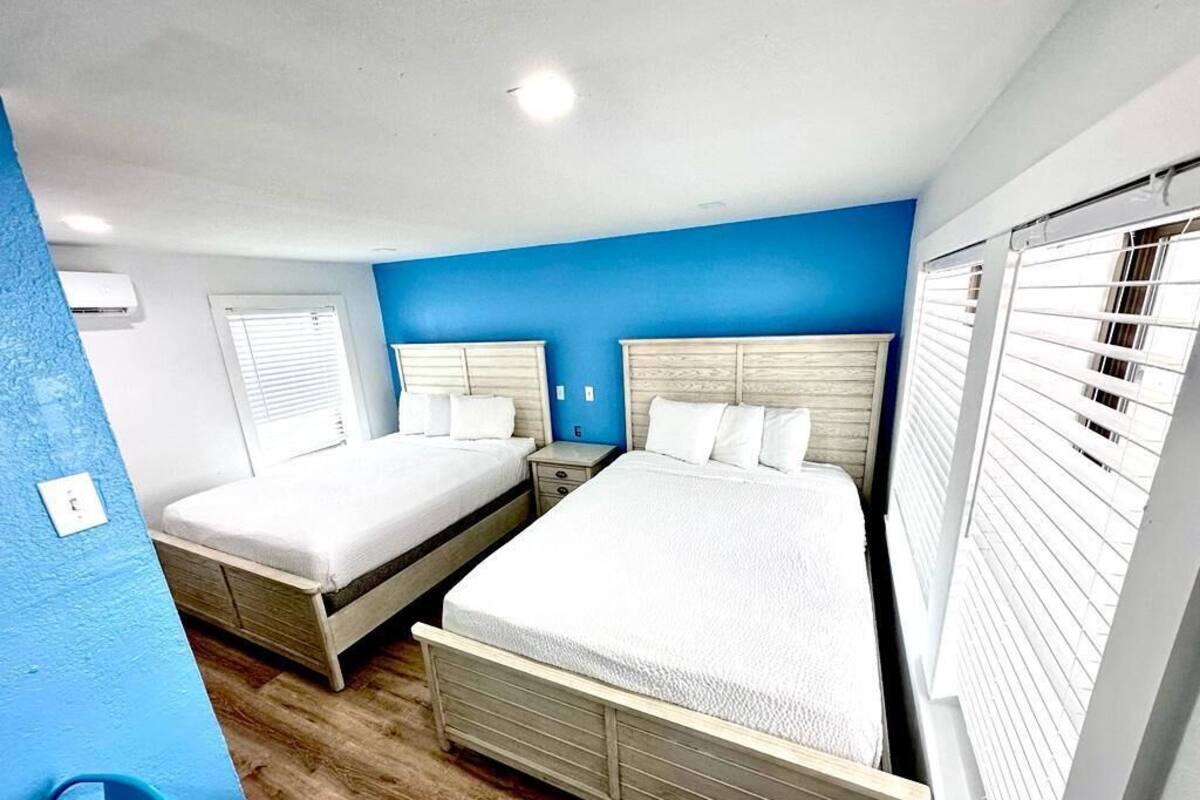 A hotel room with two beds, a blue accent wall, wooden flooring, and a window with blinds.
