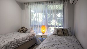 A bedroom with two beds, a nightstand with a lamp, and a window with sheer curtains. - Wellington Parade 15a, Shelly Beach, Caloundra (SHELLY BEACH)