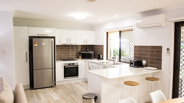 A modern kitchen with white cabinets, a stainless steel refrigerator, a microwave, and a coffee maker. There is a window with a view of greenery outside. - Wellington Parade 15a, Shelly Beach, Caloundra (SHELLY BEACH)