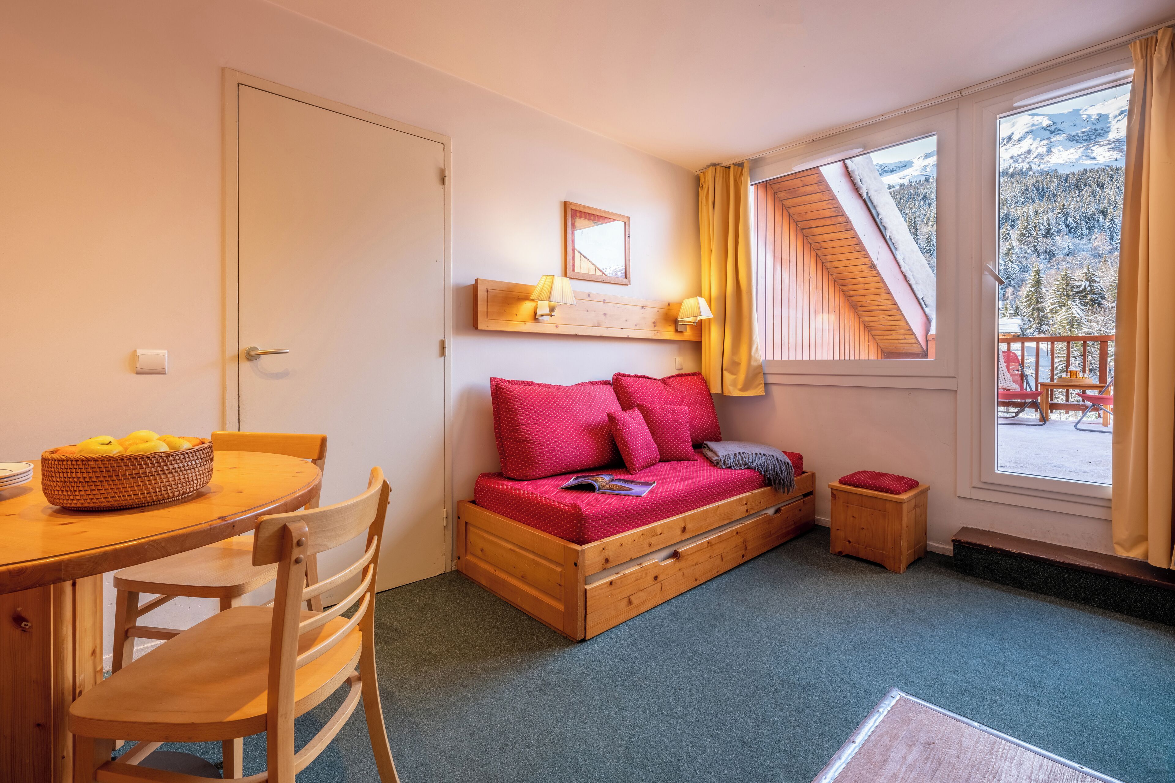 A cozy room with a wooden bed, a small table with a bowl of fruit, and a window with a view of a snowy mountain.