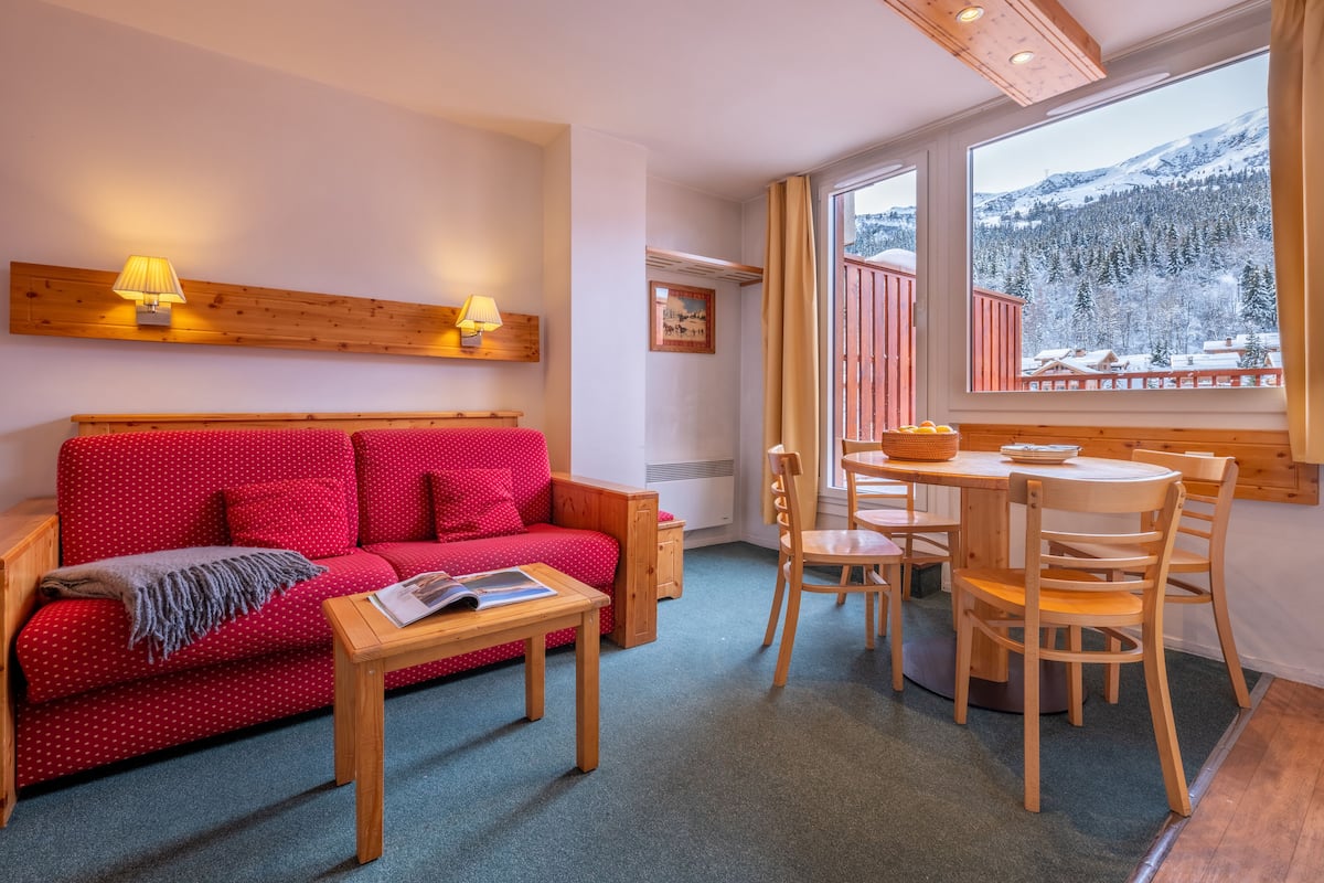 Un cuarto acogedor con un sofá rojo de lunares, una mesa de madera y sillas, una ventana con vistas a una montaña nevada y un techo de madera.
