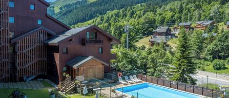 Un bâtiment en bois avec une piscine, entouré d’une zone délimitée par une clôture, où se trouvent des chaises longues et des parasols.