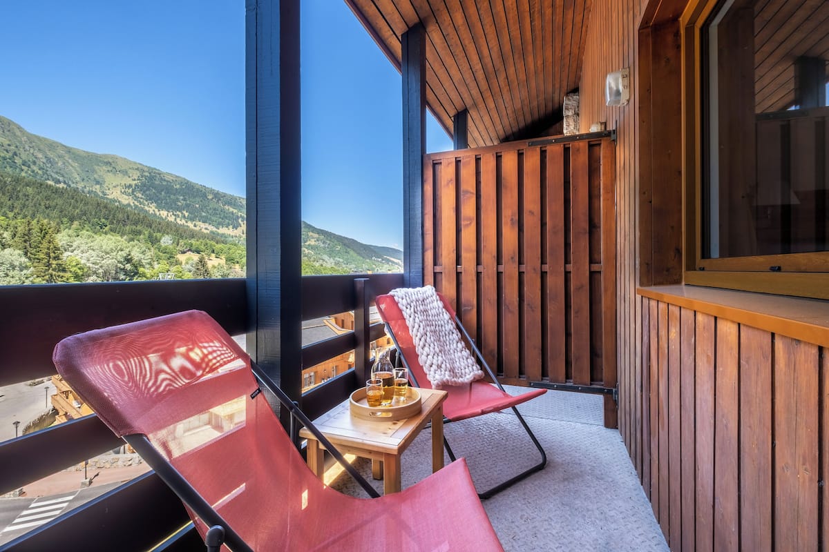 Un balcón con dos sillas rojas, una mesa pequeña con una bandeja de bebidas y vistas a un paisaje montañoso.
