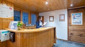 A wooden reception desk with a woman holding a document, a weather forecast sign, and a framed poster on the wall. -  Résidence Pierre & Vacances Résidence Le Peillon (Les Allues)