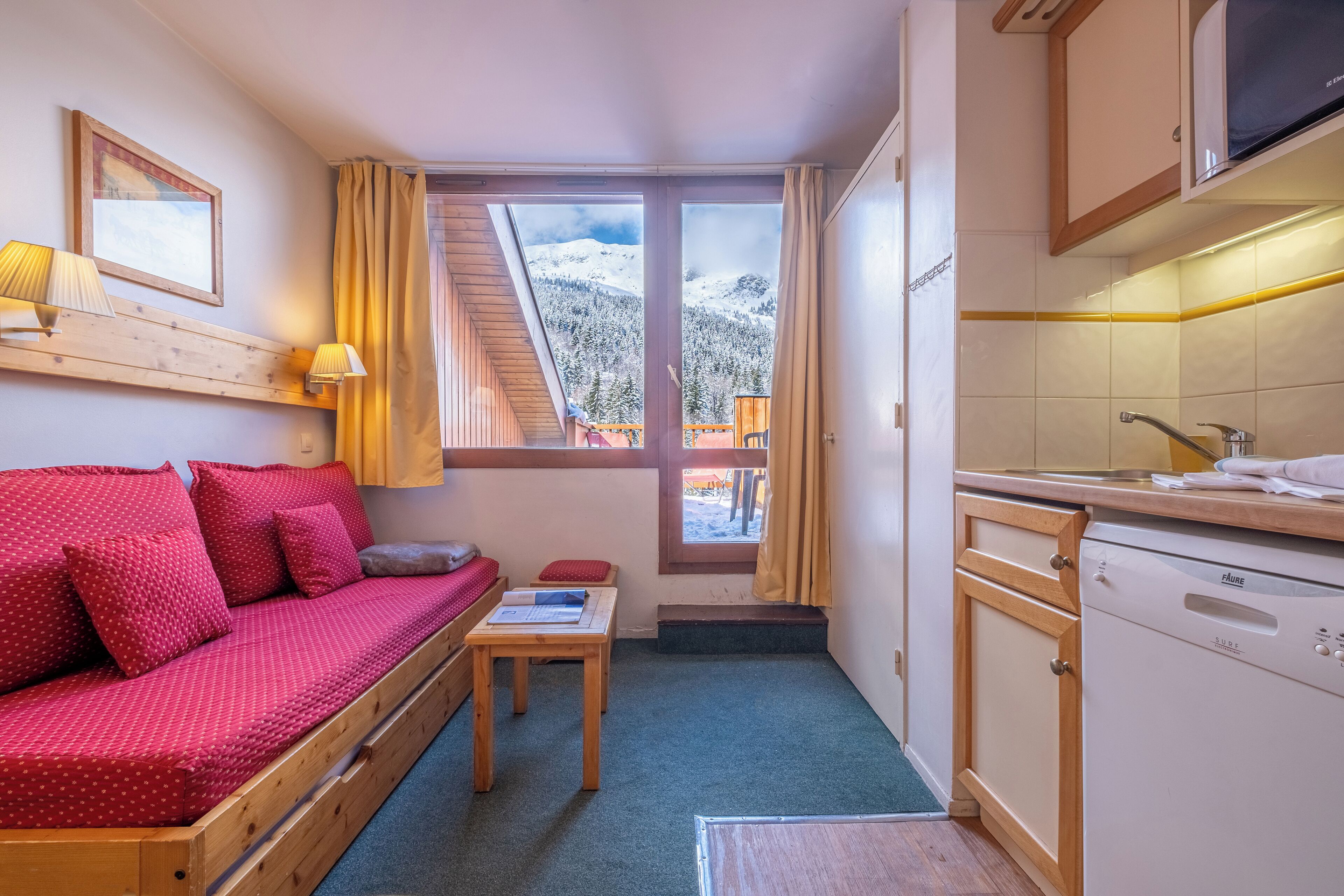 A compact room with a wooden sofa, a small table, a window with a view of snow-covered mountains, and a kitchen area with a dishwasher.