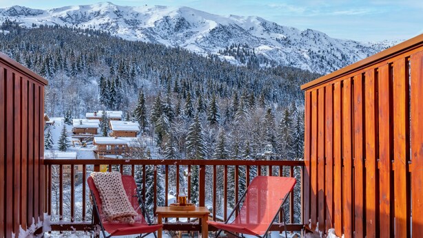A balcony with two red chairs and a small table, overlooking a snowy mountain landscape with cabins. -  Résidence Pierre & Vacances Résidence Le Peillon (Les Allues)