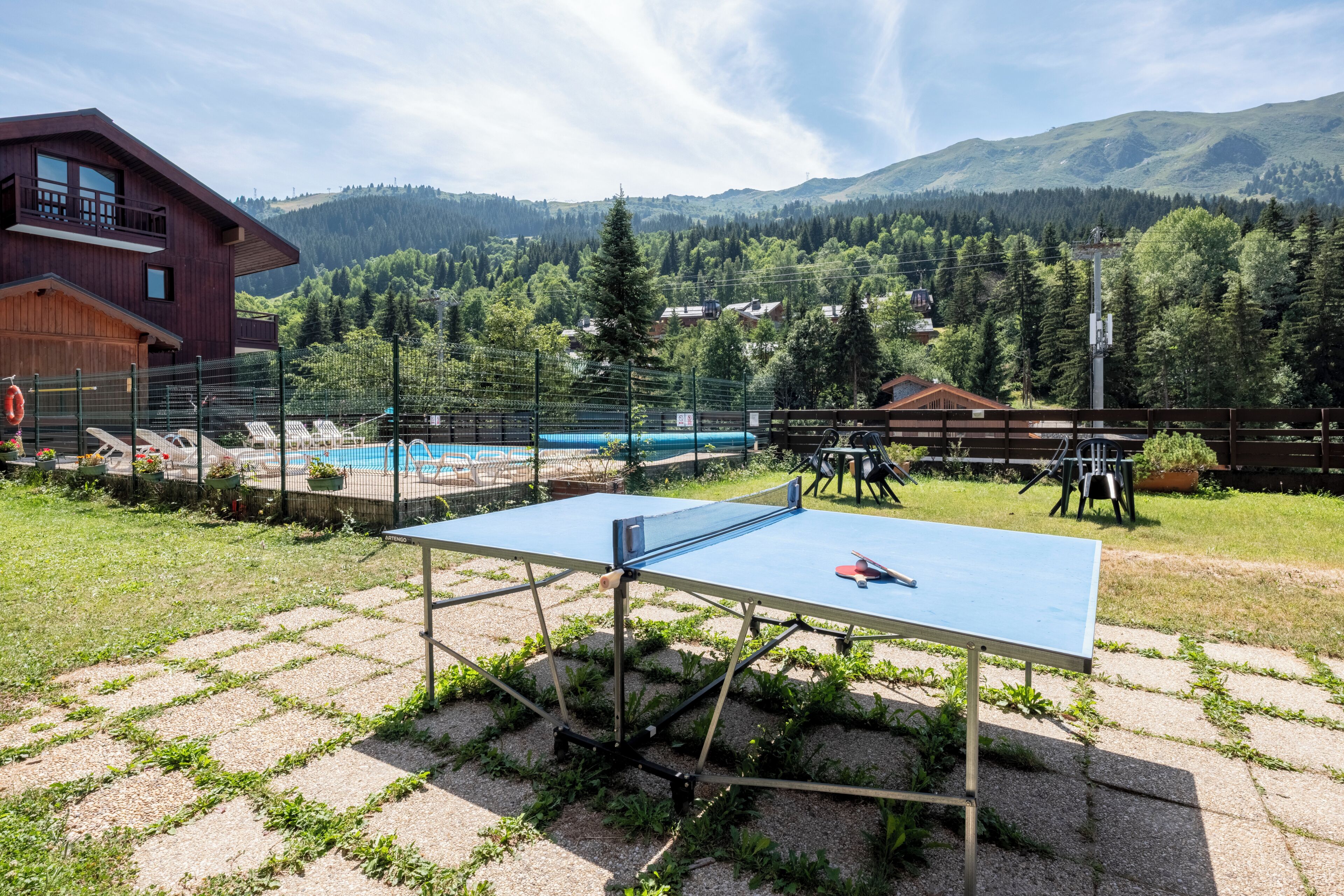 Mesa de ping pong al aire libre con vista a la montaña, rodeada de una zona de piscina y un edificio de madera.