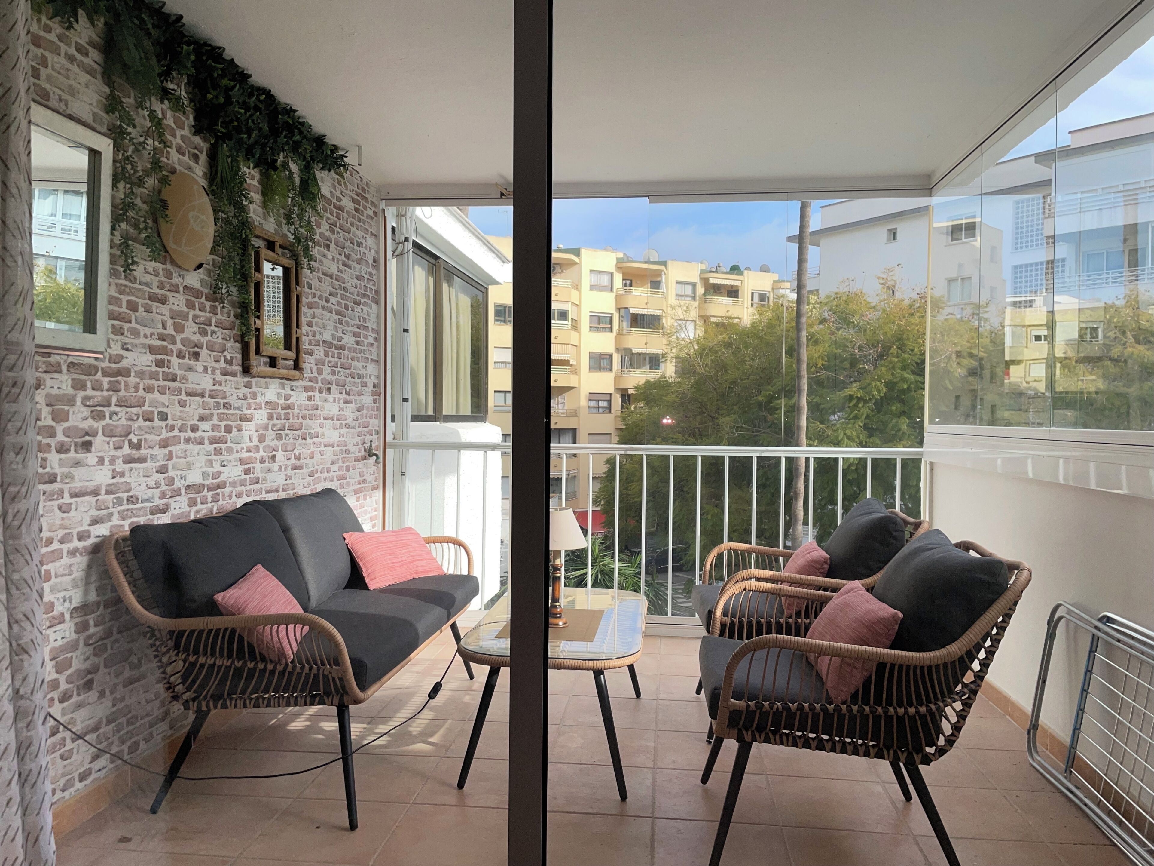 Un balcón con mobiliario de mimbre, una mesa de vidrio y vistas a edificios.