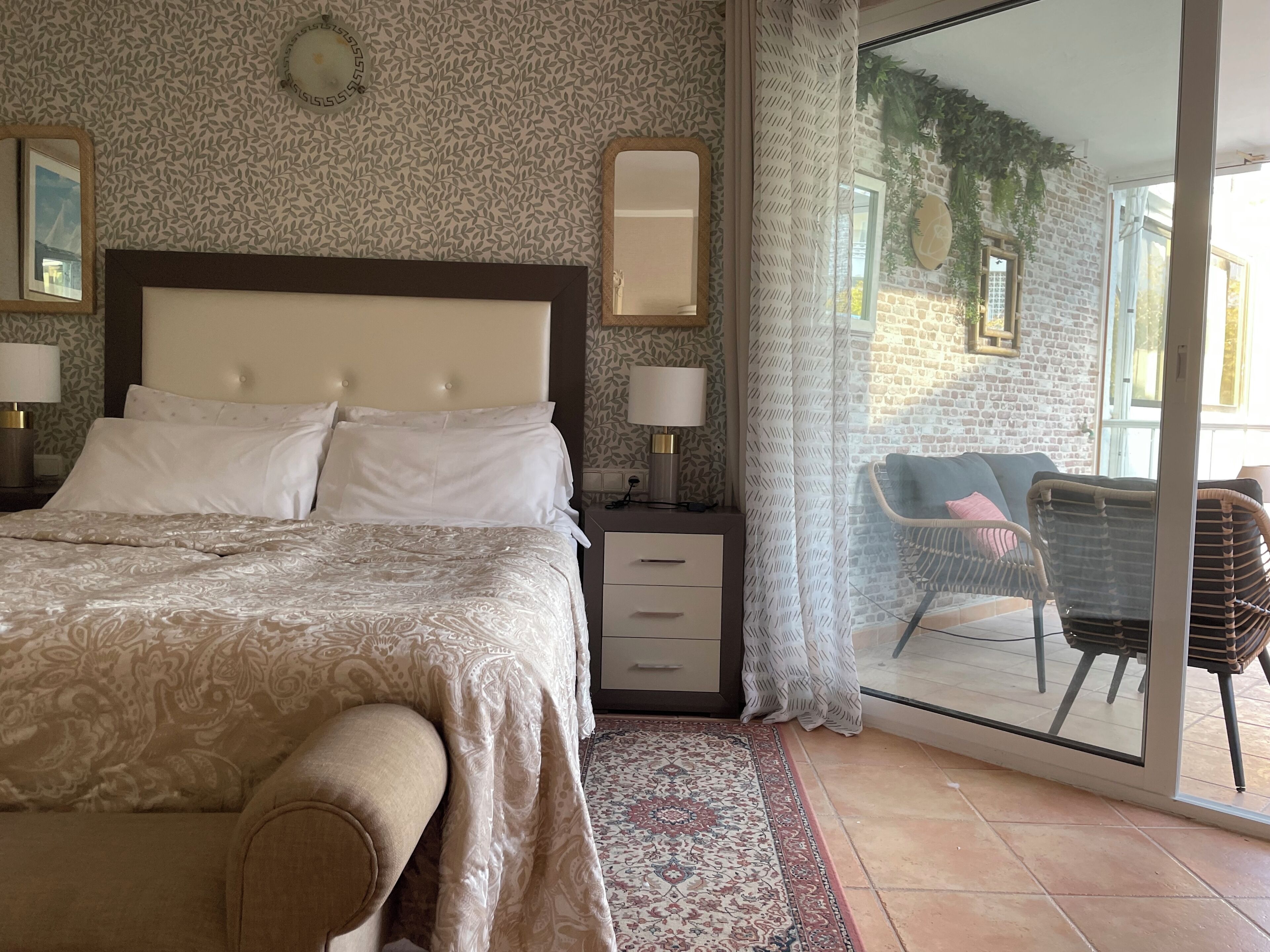 A bedroom with a bed, bedside tables, a nightstand, a patterned rug, and a view of an outdoor area with a chair and a table.