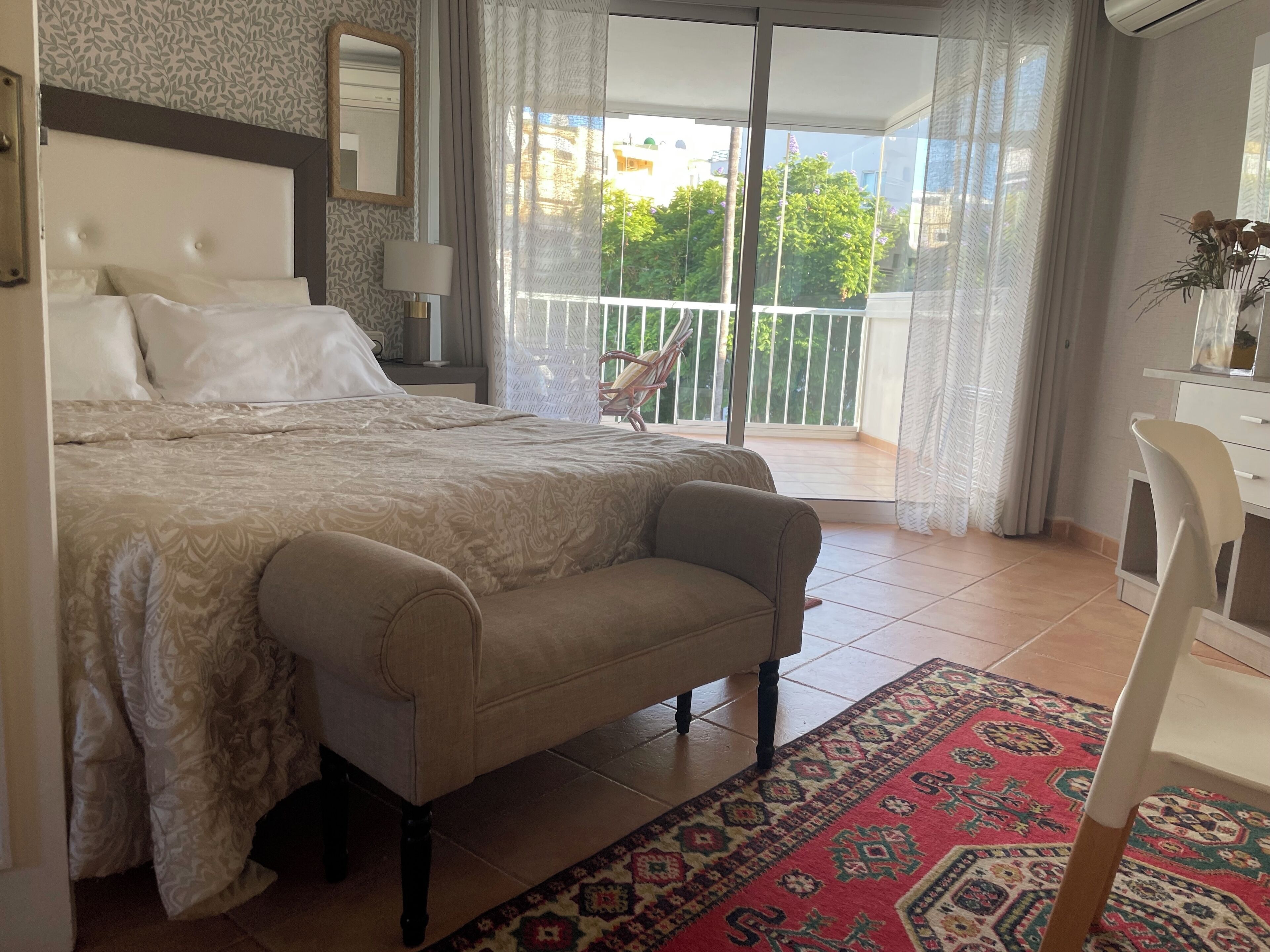 Een slaapkamer met een bed, een stoel, een nachtkastje, een lamp, een kast, een spiegel en een balkon met uitzicht op groen.