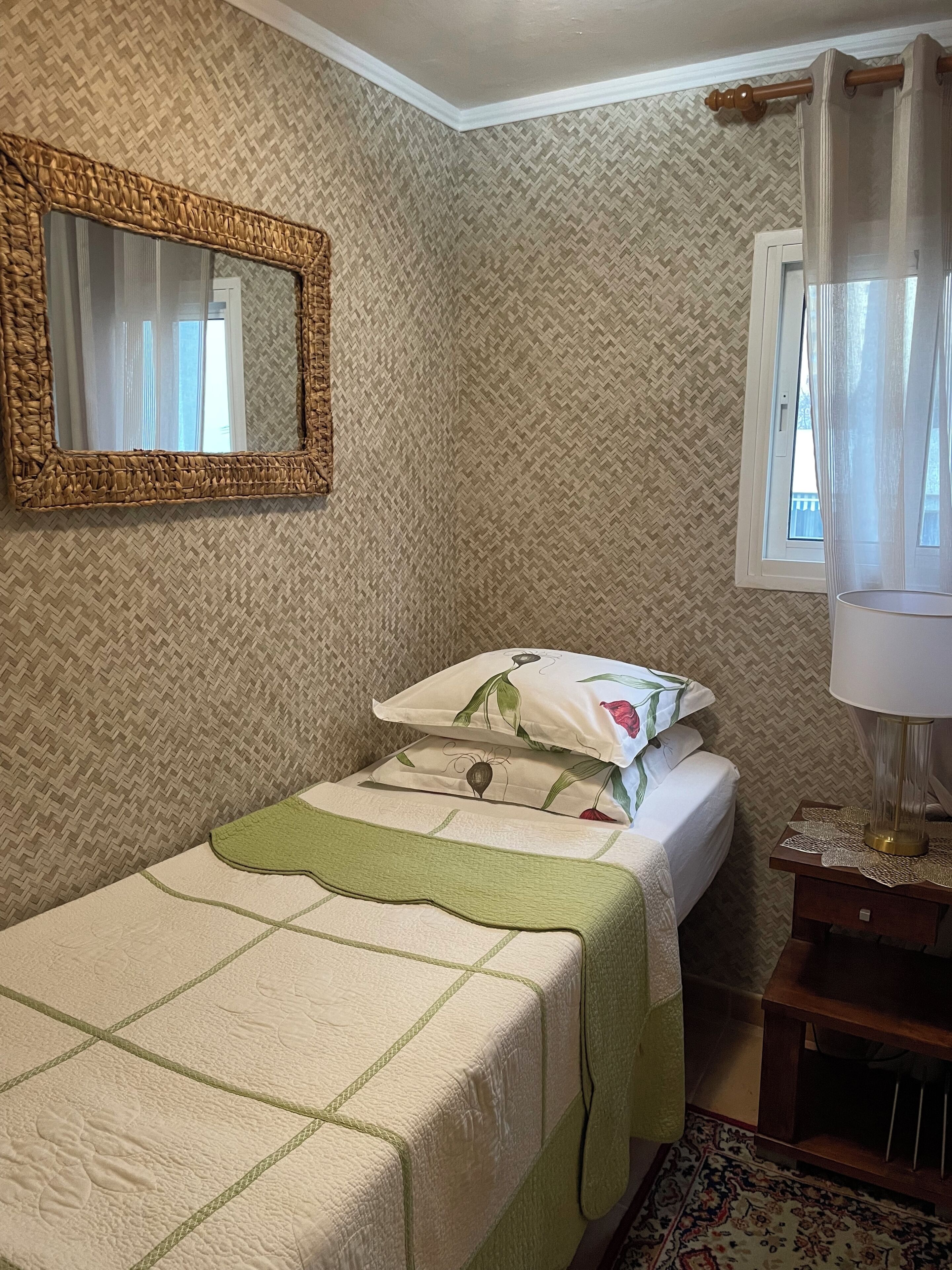 A bedroom with a bed, a bedside table, a lamp, a window with curtains, a mirror, and patterned wallpaper.