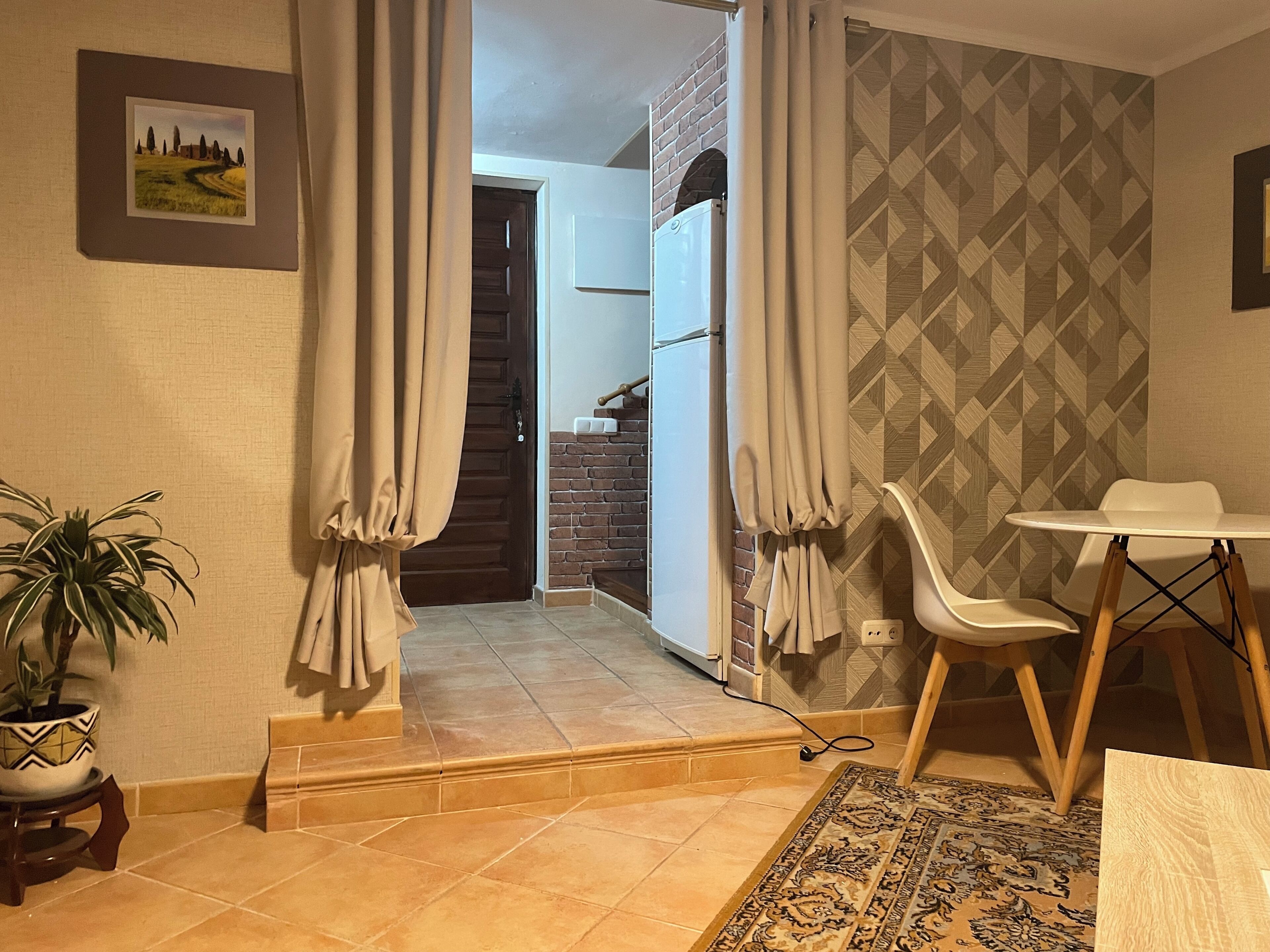 A room with a dining table, chairs, a potted plant, a patterned rug, a door with curtains, and a kitchen area in the background.