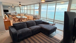 A modern living room with a sectional sofa, a dining area with a table and chairs, and large windows offering a view of the ocean. - Oceanfront Beachfront Penthouse, Isla Verde (Carolina)