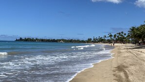 A beach with clear blue water, a sandy shore, and palm trees. - Oceanfront Beachfront Penthouse, Isla Verde (Carolina)
