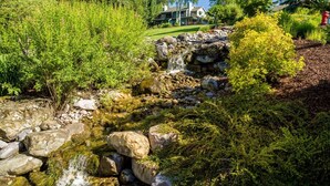 A natural water feature with rocks and greenery, a well-maintained lawn, and a house in the background. - Mountain Harbor #202 2 Bedroom Condo (Whitefish)