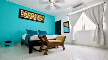 A bedroom with a bed, a wooden chair, a ceiling fan, and artwork on the walls.