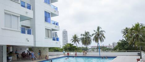 A swimming pool area with a building, palm trees, and a view of a tall building in the background.