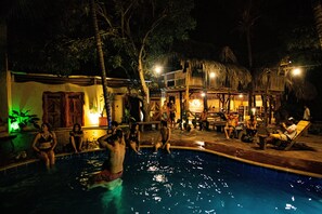 A nighttime pool area with people relaxing and socializing, surrounded by thatched-roof structures and palm trees. - Kanta Sana (Dibulla)